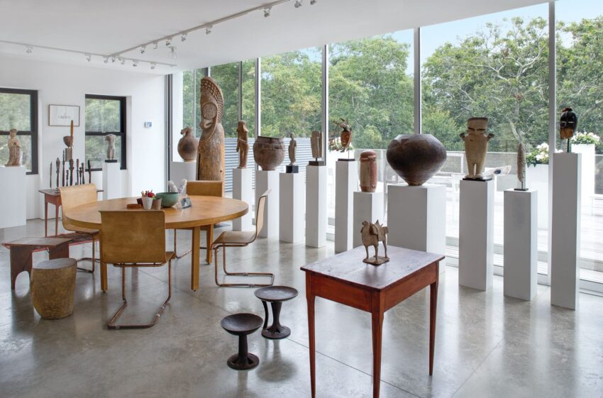 Gallery interior with diverse sculptures and pottery on pedestals, wooden tables and stools, large windows with garden view.