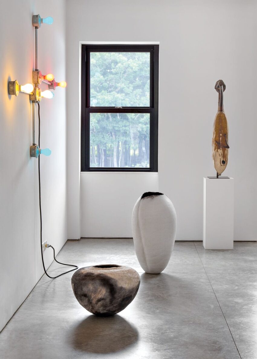 Modern art gallery with sculptures, colorful wall lights, and a large window with a view of trees.