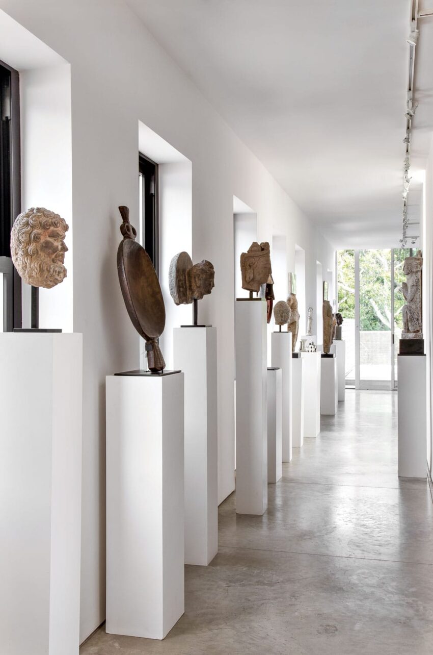 Art gallery hallway with sculptures on white pedestals, featuring various busts and a shield, lit by natural light from windows.