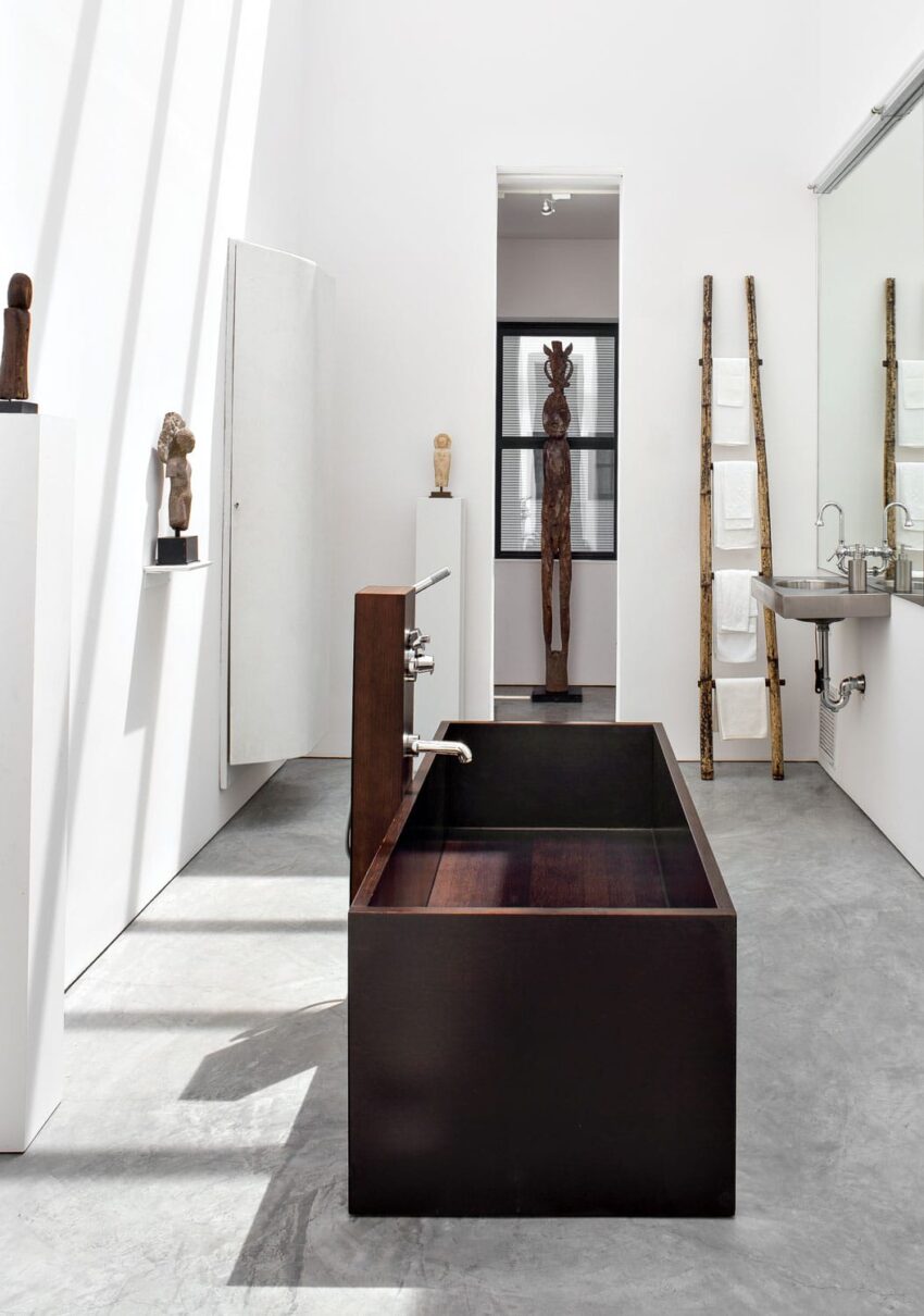 Modern minimalist bathroom with a dark wooden bathtub, sculptures, ladder towel rack, and sleek fixtures.