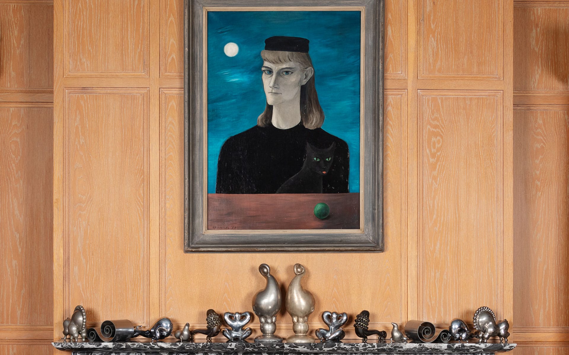 Framed portrait of a woman and a cat on a wooden wall above a shelf with decorative metal sculptures.