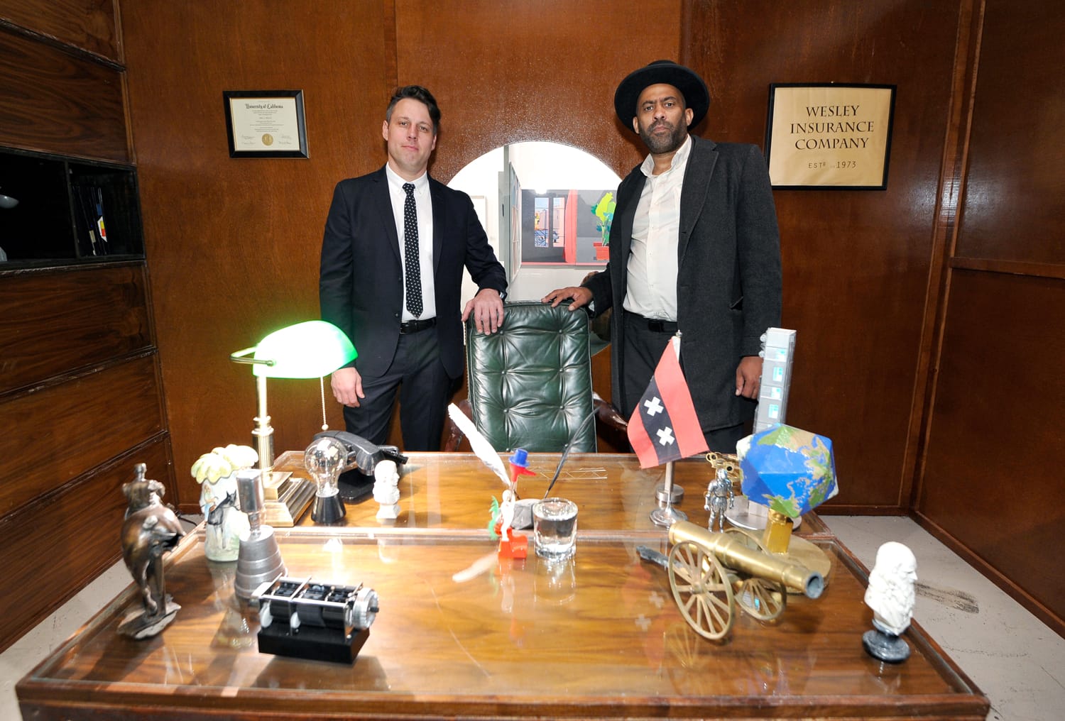 Two men standing in a vintage office behind a wooden desk with various decorative items and a Wesley Insurance Company sign.