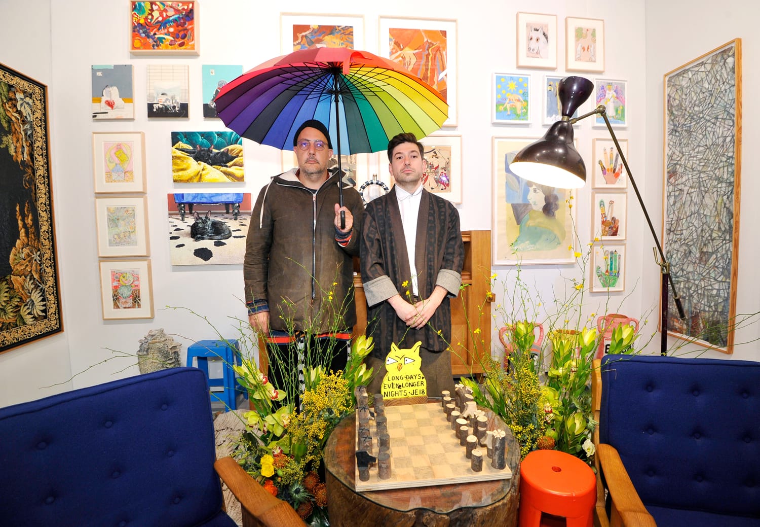 Two people stand indoors under a colorful umbrella surrounded by art, flowers, and a chessboard in a vibrant setting.