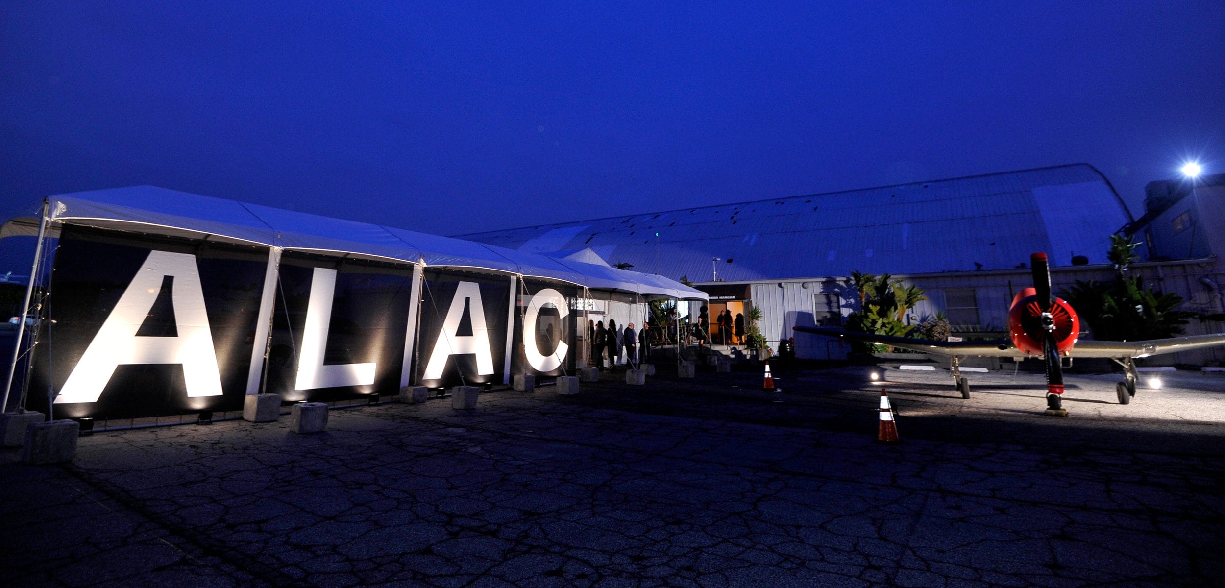 Large illuminated "ALAC" sign at an outdoor event with a vintage airplane nearby during nighttime.