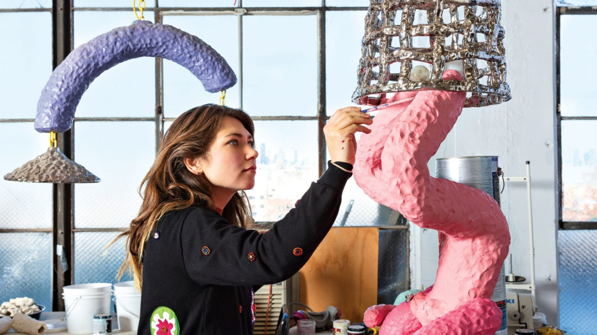 Artist working on abstract sculptures in a studio with large windows and city skyline in the background.
