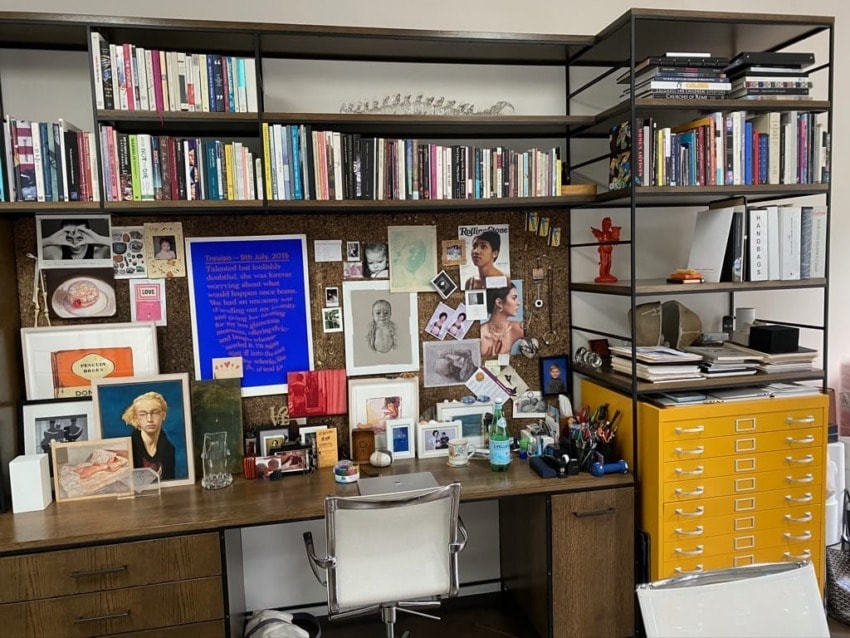 Cluttered home office desk with books, art, and personal items on shelves and corkboard.