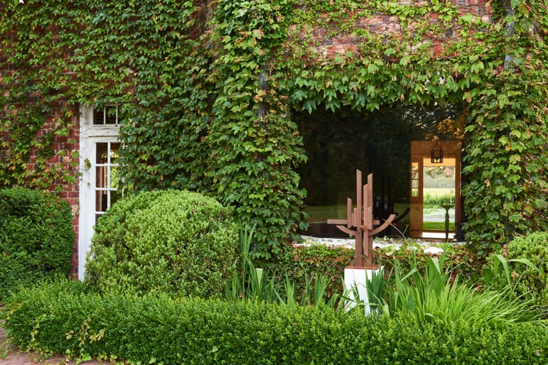 Brick house covered in ivy with a large window and sculpted greenery in the garden.