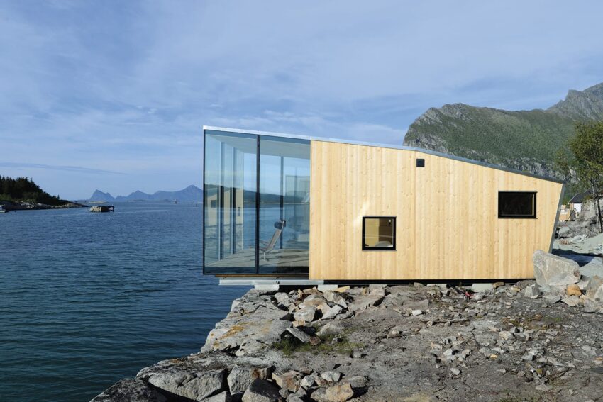 Modern wooden cabin with large glass window overlooking a rocky shoreline and calm sea with mountains in the background.