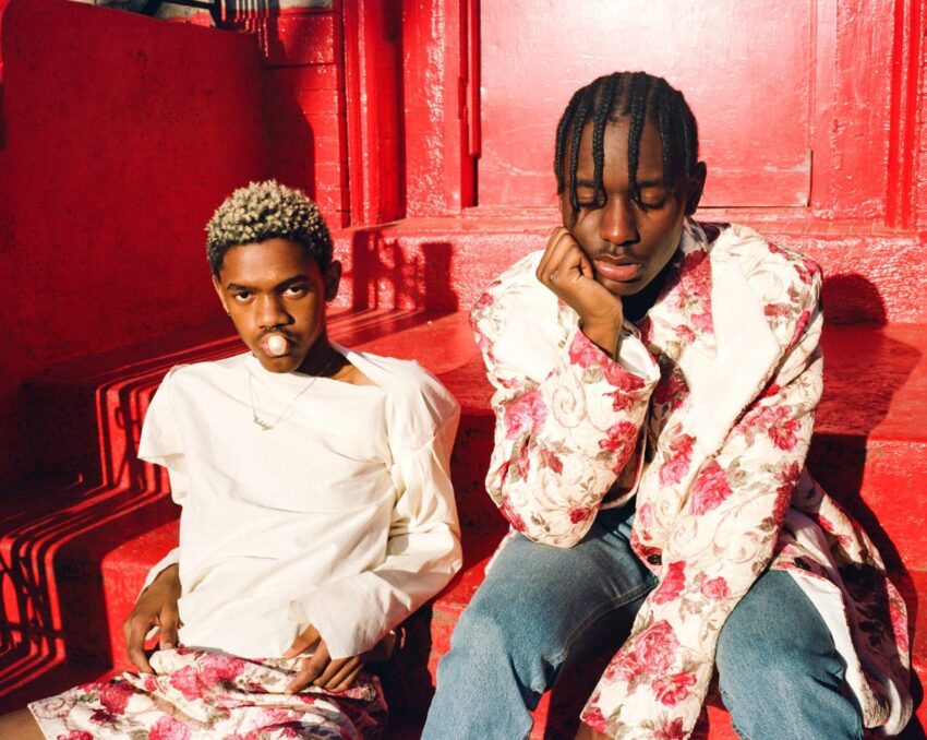 Two stylish individuals in floral outfits sit on red steps, one blowing a bubble gum, the other with hand on chin.