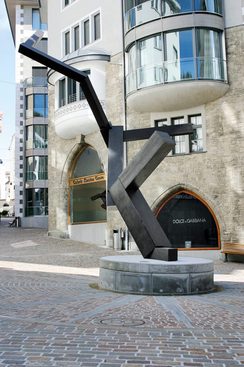 Modern abstract metal sculpture in urban plaza with nearby contemporary buildings and shops in the background
