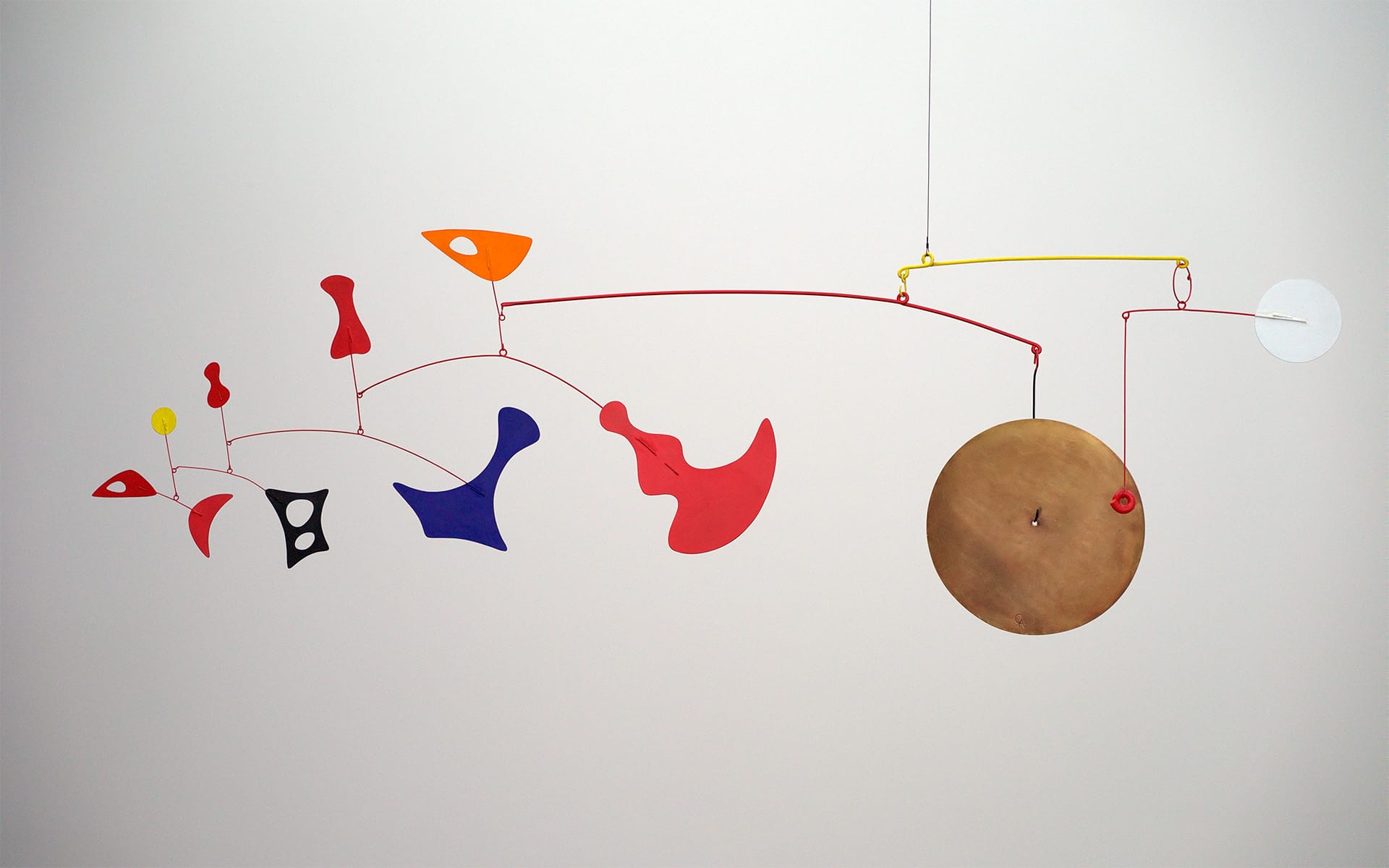 Colorful abstract mobile art sculpture with various shapes and a large bronze disc hanging against a white wall.