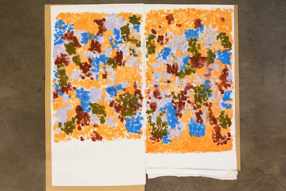 Colorful abstract painting with splashes of orange, blue, brown, and green on paper laid on a concrete floor.