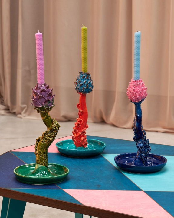 Three colorful sculpted candleholders with tall pink, coral, and blue candles on a modern geometric table.