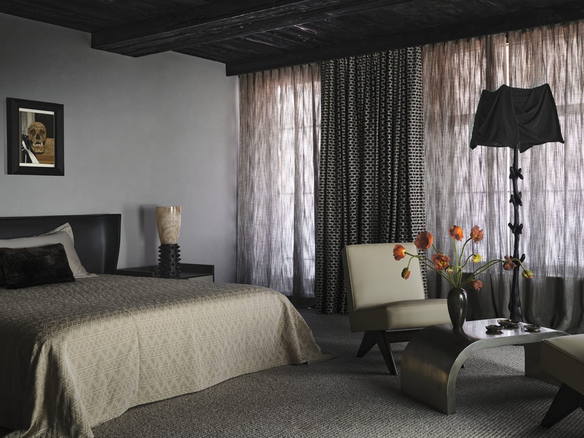 Modern bedroom with a beige bed, patterned curtains, elegant furniture, and a skull painting on the wall.