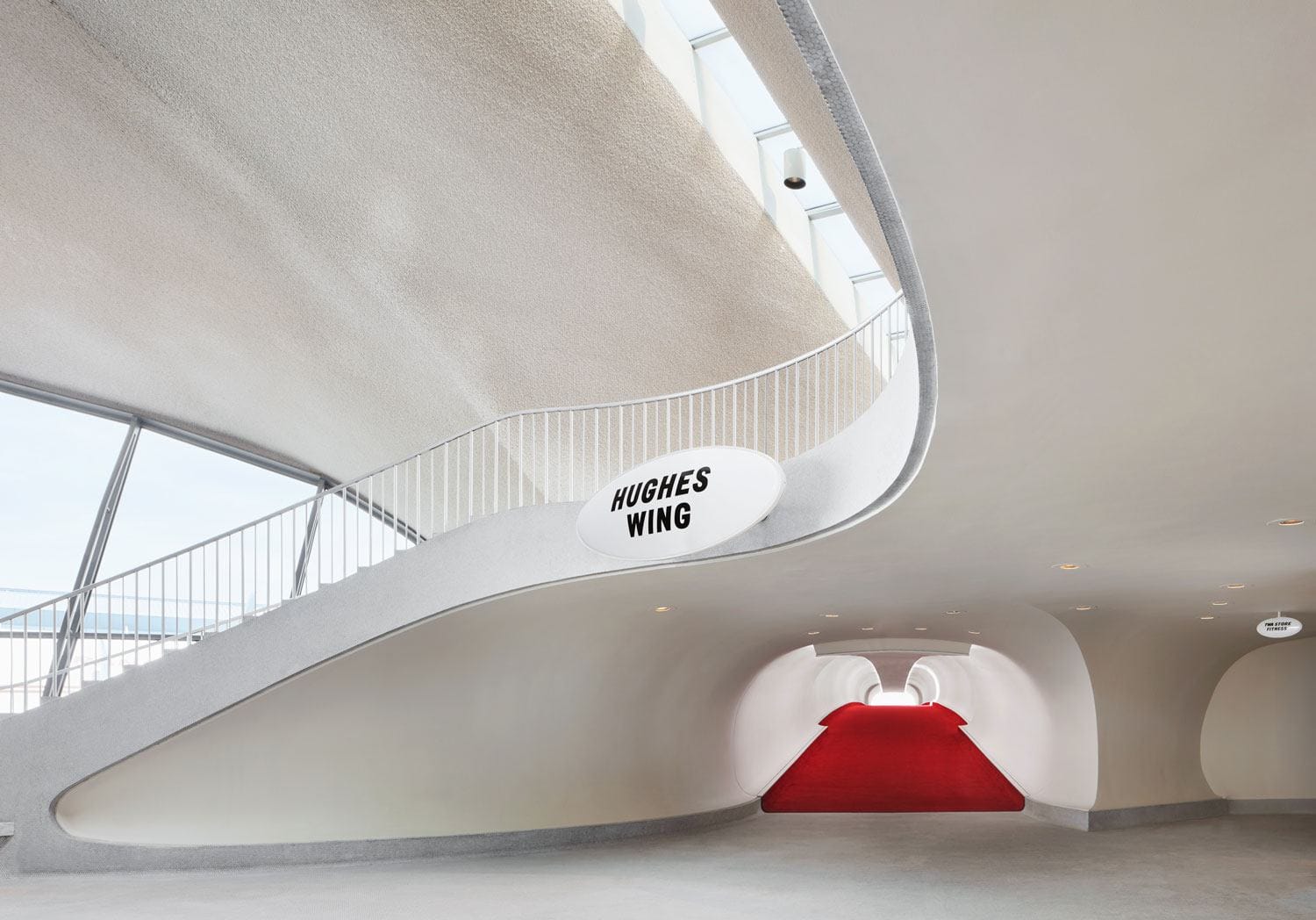 Futuristic curved staircase with a red carpet, leading to the "Hughes Wing" sign in a modern architectural setting.