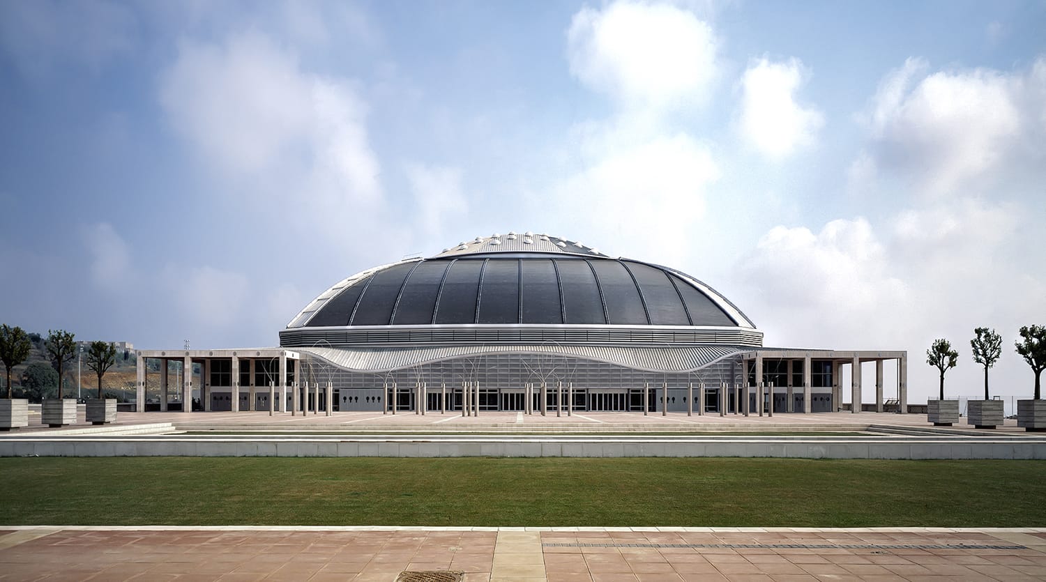 Large modern dome-shaped building with a curved roof, set against a cloudy sky and surrounded by a green lawn and trees.