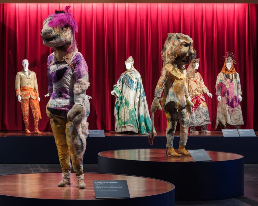 Exhibit of colorful, artistic costumes and large, unique animal figures against a red curtain backdrop.