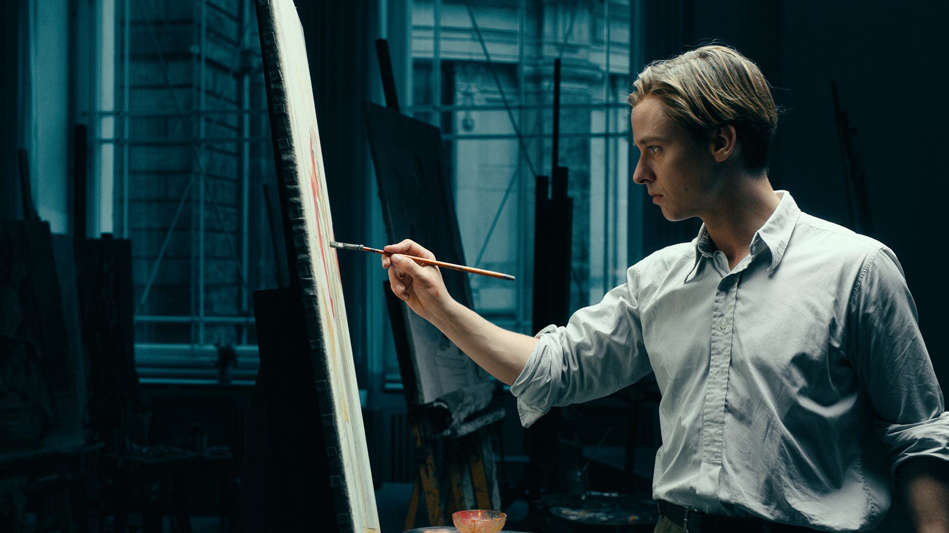 Artist painting on a large canvas in a dimly lit studio with art supplies and easels in the background