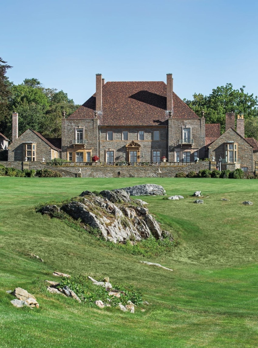 Large stone mansion with red roof surrounded by greenery and expansive lawn under clear blue sky