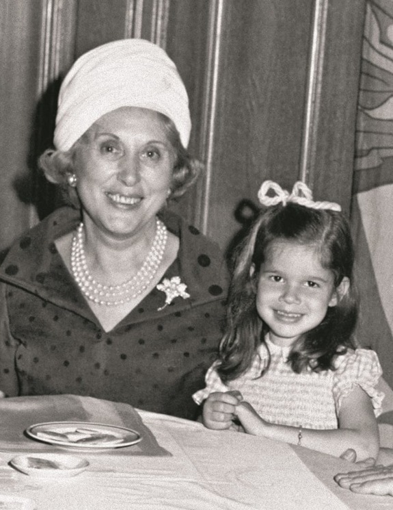 Elderly woman with a white headscarf and young girl smiling at a table with a plate and napkin in front of them.