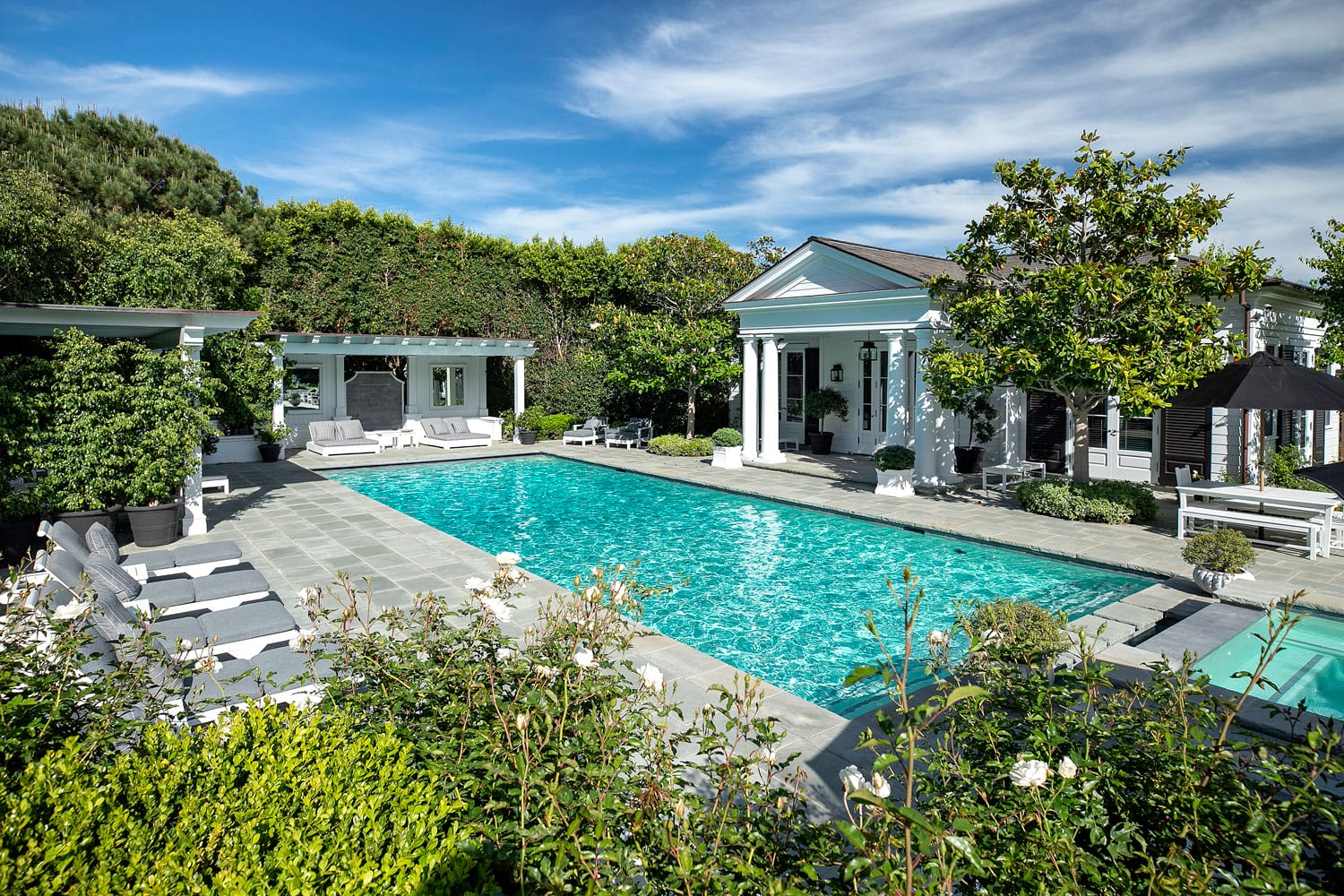 Elegant white villa with a large swimming pool surrounded by greenery and clear blue sky in the background.
