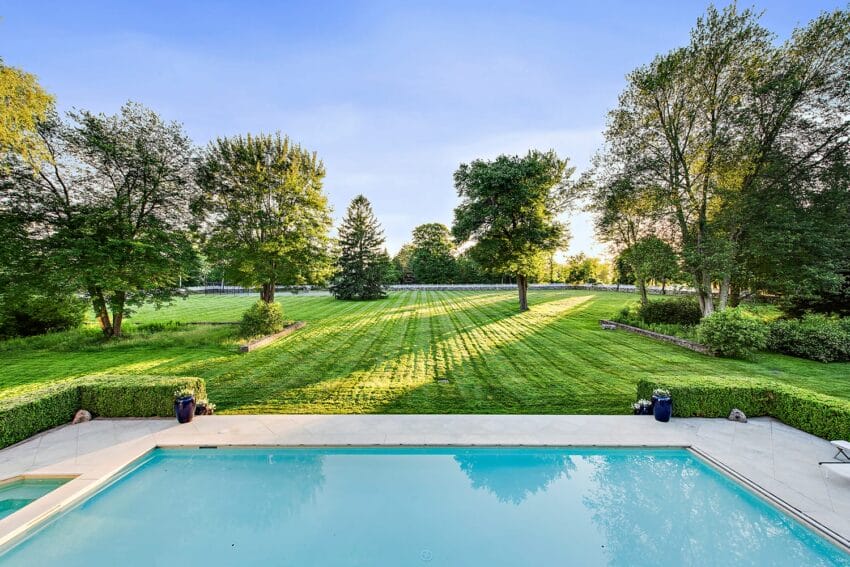 Backyard view with a swimming pool in the foreground, green lawn, lush trees, and clear blue sky in the background.