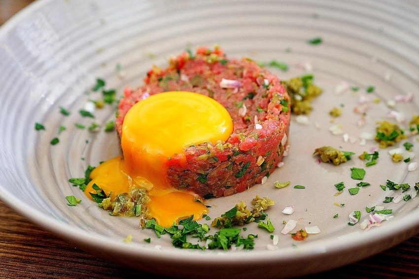 Beef tartare topped with a raw egg yolk, garnished with chopped herbs and onions on a ceramic plate