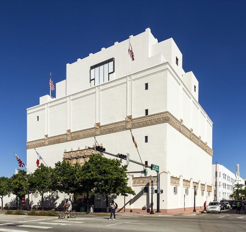 Art Deco-style building with flags, people, and bicycles on a sunny street corner under a clear blue sky.