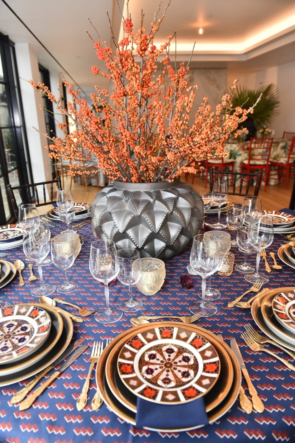 Elegant dining table setting with patterned plates, gold cutlery, glassware, and a decorative floral centerpiece.