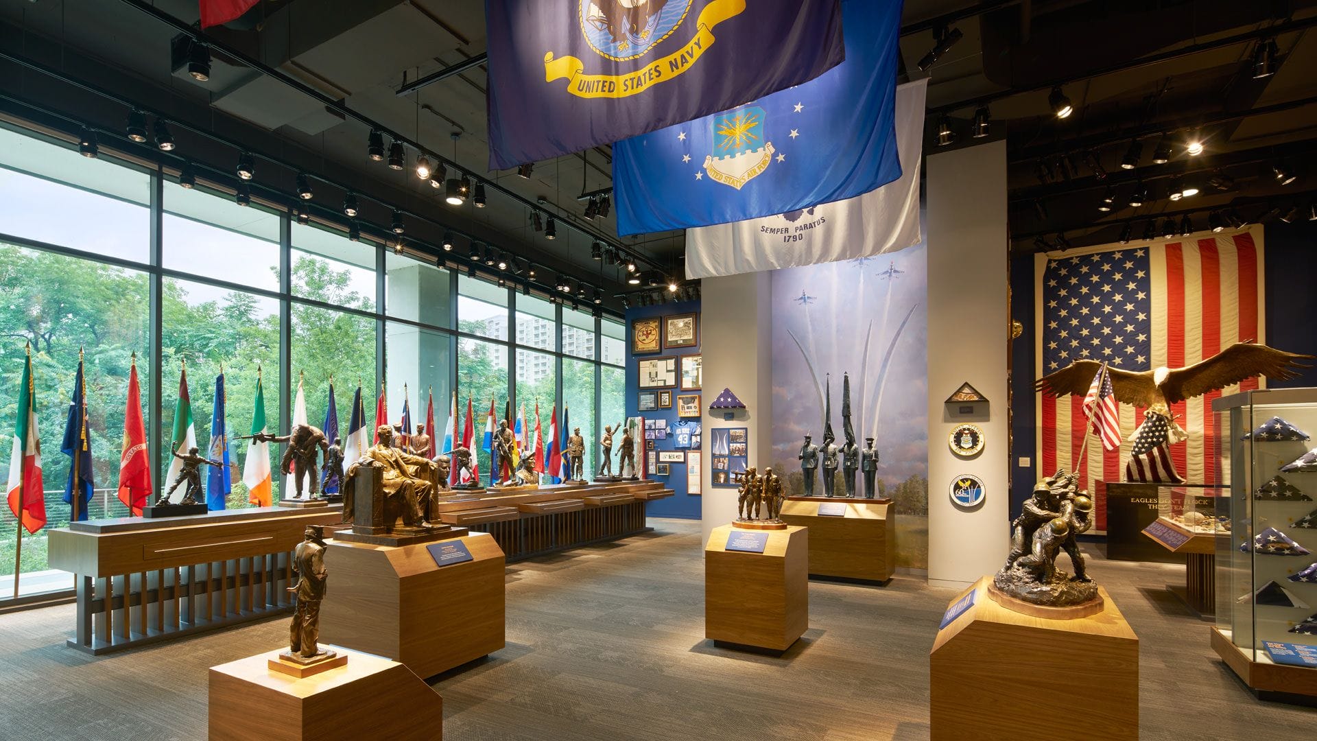 Museum exhibit room with military flags, sculptures, and large windows displaying a view of trees outside.
