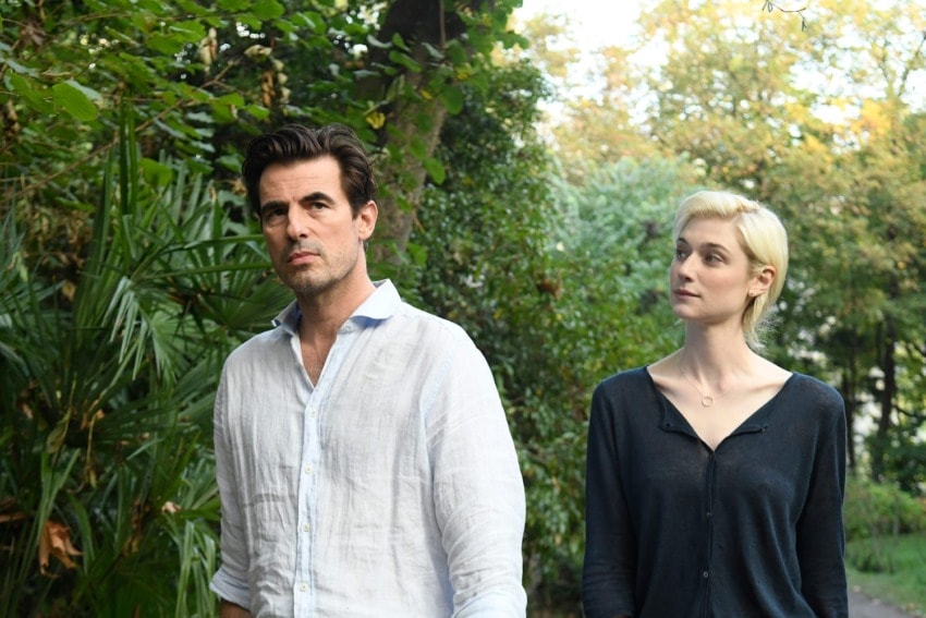 Man and woman walking through a lush garden with tall trees and green foliage in the background
