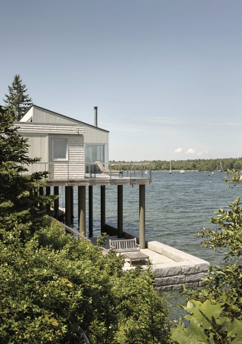 Modern lakeside cabin on stilts with wooden deck, surrounded by lush greenery and overlooking calm waters and distant sailboats.