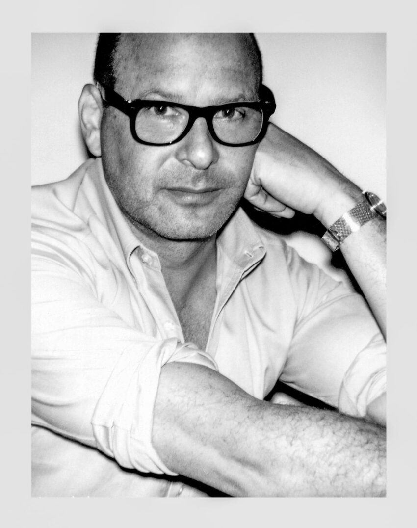 Man with glasses sitting, wearing a rolled-up shirt and bracelet, looking at the camera with a neutral expression.