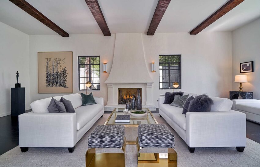 Modern living room with white sofas, a central coffee table, a fireplace, wooden ceiling beams, and wall art.