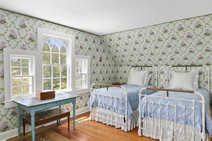 Vintage bedroom with floral wallpaper, two white metal twin beds, blue bedding, arched window, and a wooden floor.