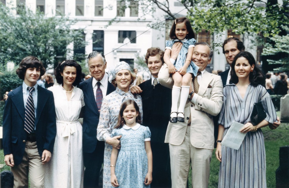 A family posing together outdoors in formal attire with trees and buildings in the background.