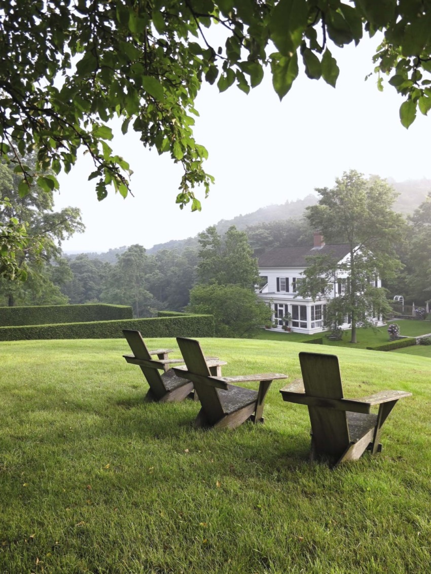 Three wooden chairs on a lush green hill overlooking a white house with trees in the background.