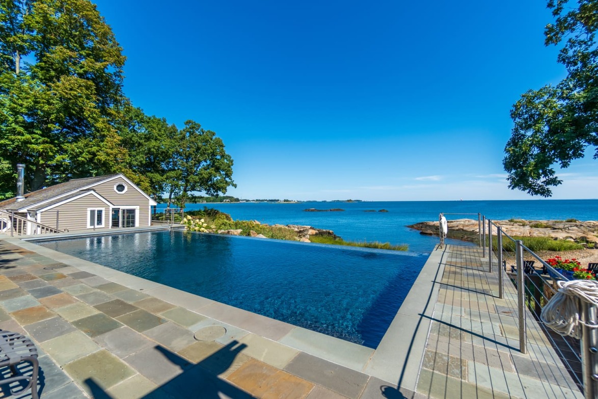 Infinity pool overlooking a serene ocean view with a small cottage and lush trees on a sunny day.