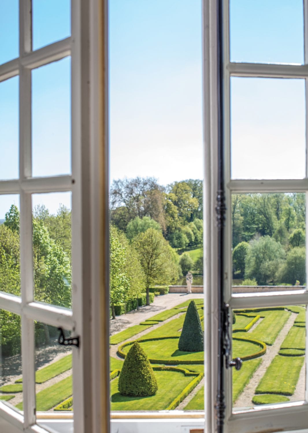 View through a window of a manicured garden with geometric hedges and trees under a clear blue sky.