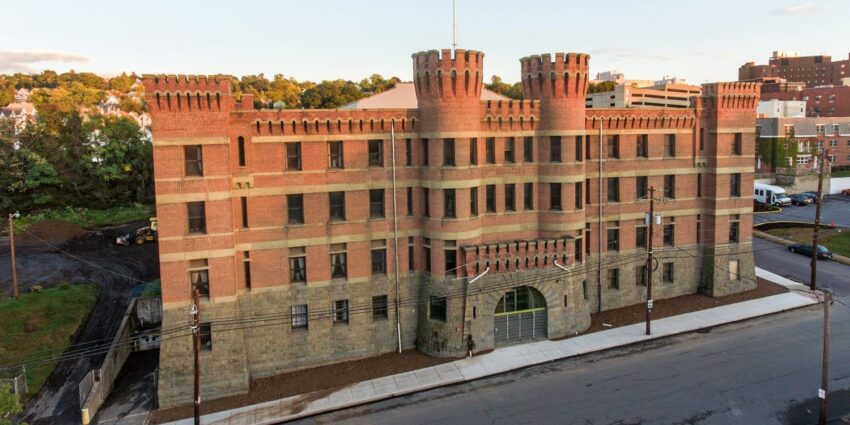 Historic brick building with turrets and arched entry, surrounded by trees and urban landscape under a clear sky.