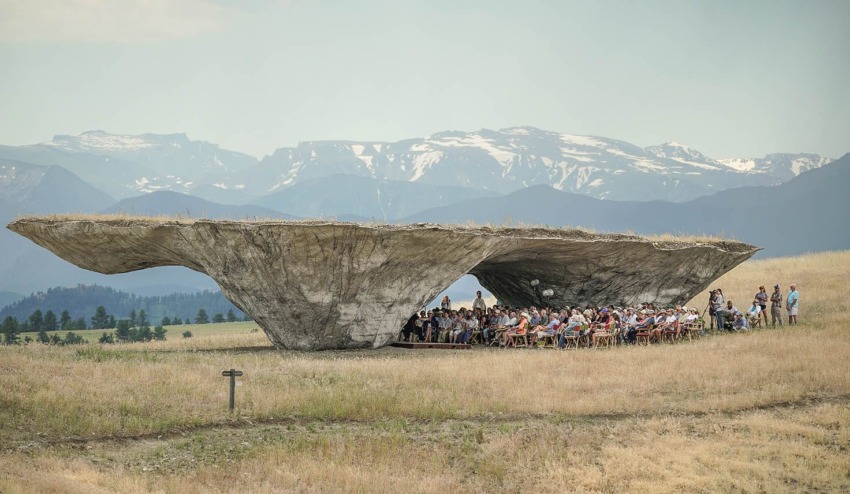 Outdoor gathering under unique, large concrete structure in a grassy field with distant mountains in the background.