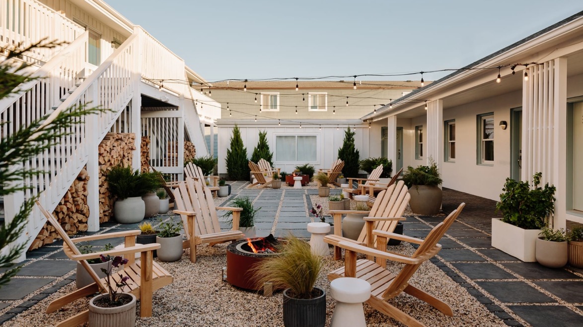 Courtyard with Adirondack chairs around a fire pit, string lights above, surrounded by potted plants and white buildings.