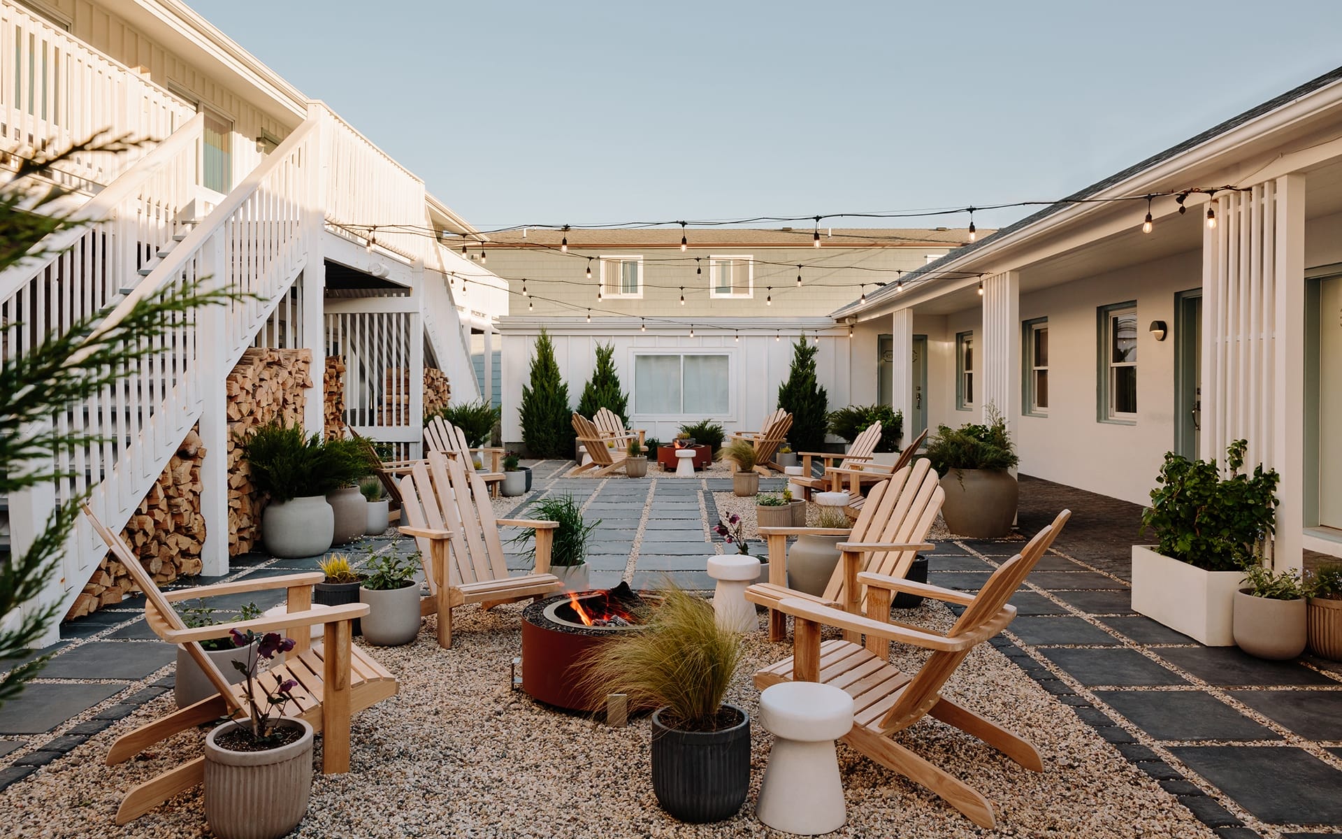 Courtyard with Adirondack chairs around a fire pit, string lights above, surrounded by potted plants and white buildings.