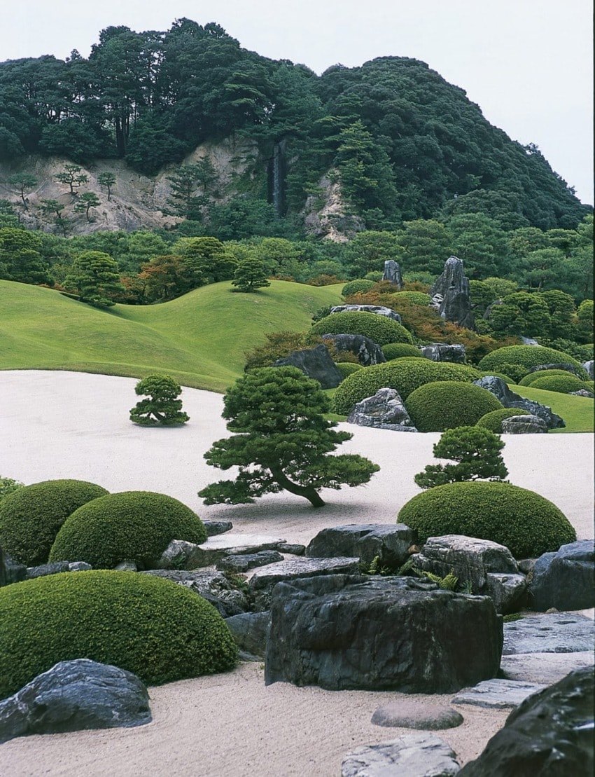 Japanese garden landscape with manicured shrubs, rock formations, and lush hills in the background.