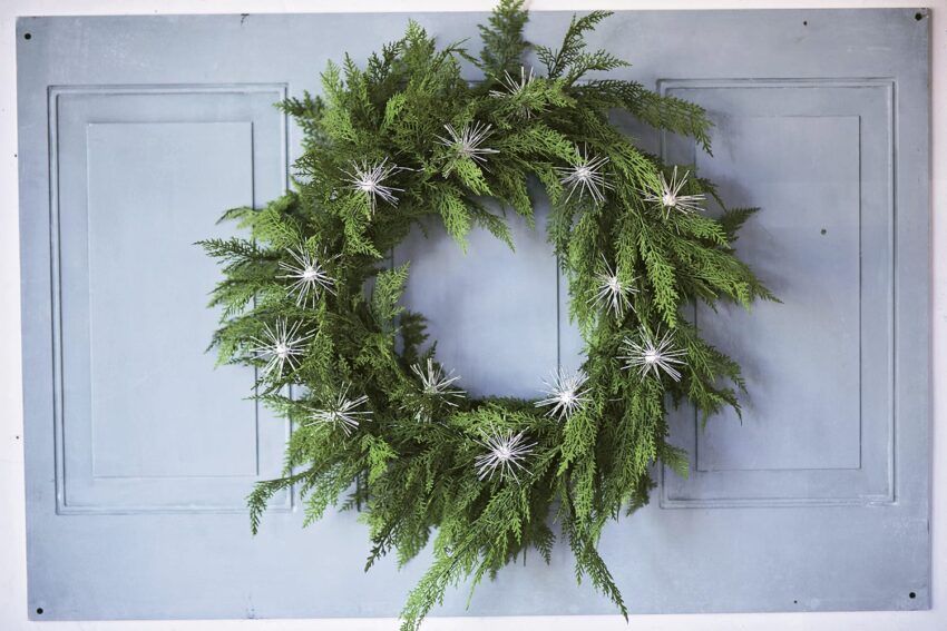 Green wreath with white star-shaped decorations on a light blue wooden panel background.