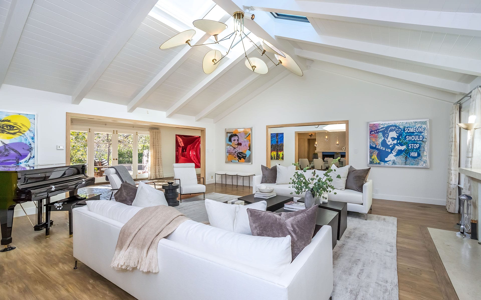 Modern living room with white sofas, art on walls, and large windows. Abundant natural light and elegant chandelier overhead.