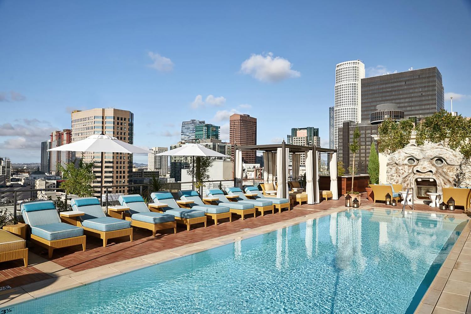 Rooftop pool with lounge chairs and umbrellas overlooking a city skyline under a clear blue sky.