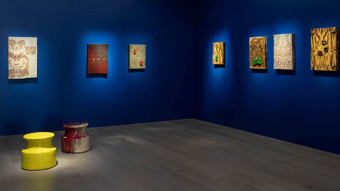Art gallery with paintings on a deep blue wall and two colorful stools on a wooden floor.