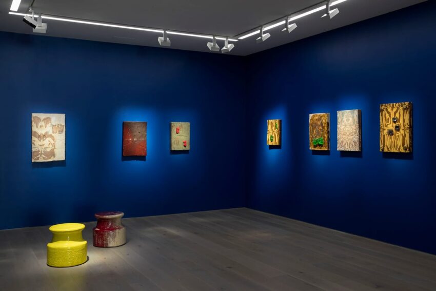 Art gallery room with blue walls, displaying various paintings and two colorful stools on the wooden floor.