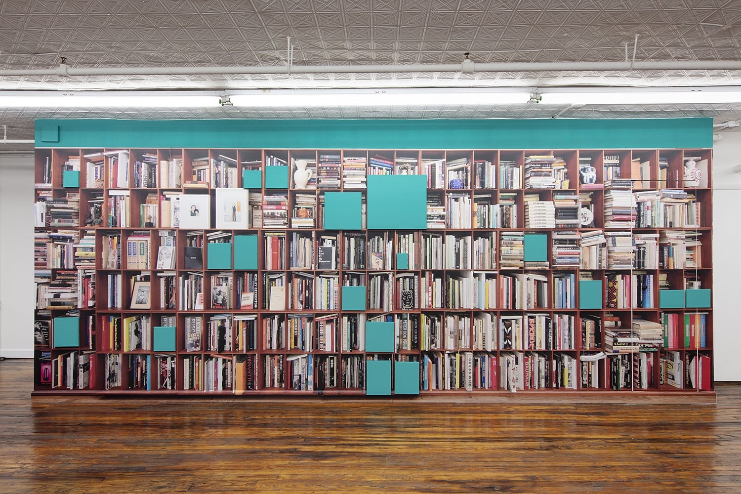 Wall-sized bookshelf with various books and teal compartments in a room with wooden flooring and decorative ceiling tiles.